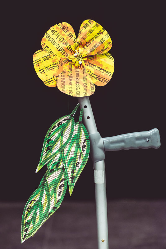 Photo of a medical grey crutch decorated with a large yellow paper flower cut from text and four leaves, again from paper with text on it. Hand written words 'relief, releaf' on the leaves.
