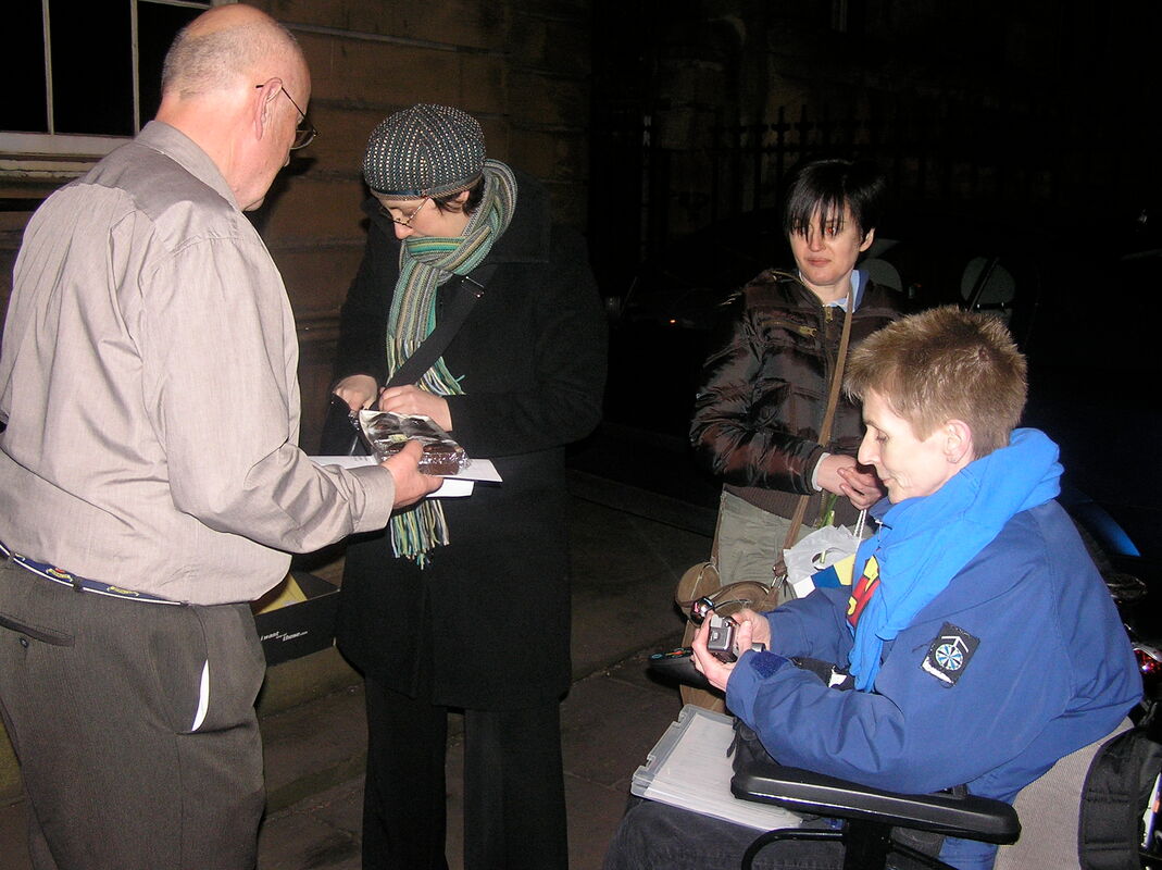 Image Description - four people outside the Lit and Phil Society in Newcastle Upon Tyne in the evening. Pauline Heath and Lindsay Carter are in the image. Lindsay is wearing a blue padded coat with a superman logo tee shirt over the top. She is seated in her powerchair. Pauline has short dark hair and is wearing grey trousers and a padded black coat. She is holding a bag of daffodils. A woman in her thirties is giving cakes and a copy of Lindsay's poem to a bald man in his fifties. He is wearing a brown shirt and trousers. 