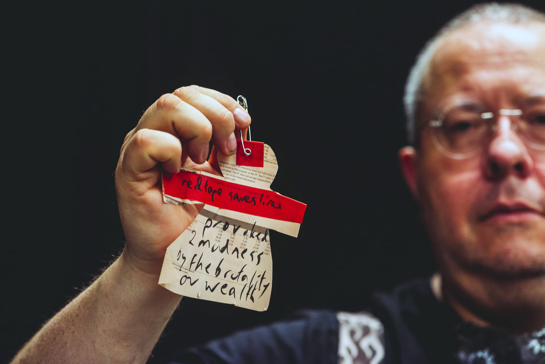 An image of gobscure, a middle aged man with a shaved head and silver rimmed round glasses, holding a paper doll cut from book text and red tape on it. Wording on the paper doll says, 'red tape saves lives' and 'provoked 2 madness by the brutality of wealth