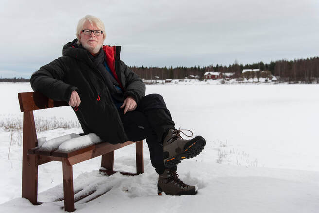 Image description. Aidan Moesby seated on a wooden bench outdoors in the snow. In the background is a pine forest and some large redbrick houses. Aidan is white man in his forties wearing dark rimmed glasses. He is well wrapped up in a padded black coat, black scarf, trousers and walking boots. 