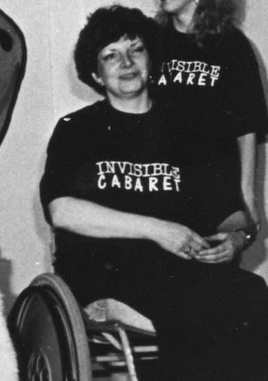 Image description - Paula Greenwell in an archive photograph of Invisible Cabaret. She is pictured on stage seated in her wheelchair. A woman in her thirties, she has short curled brown hair and is wearing an Invisible cabaret tee-shirt. Paula is smiling and holding her hands together in front of her. 