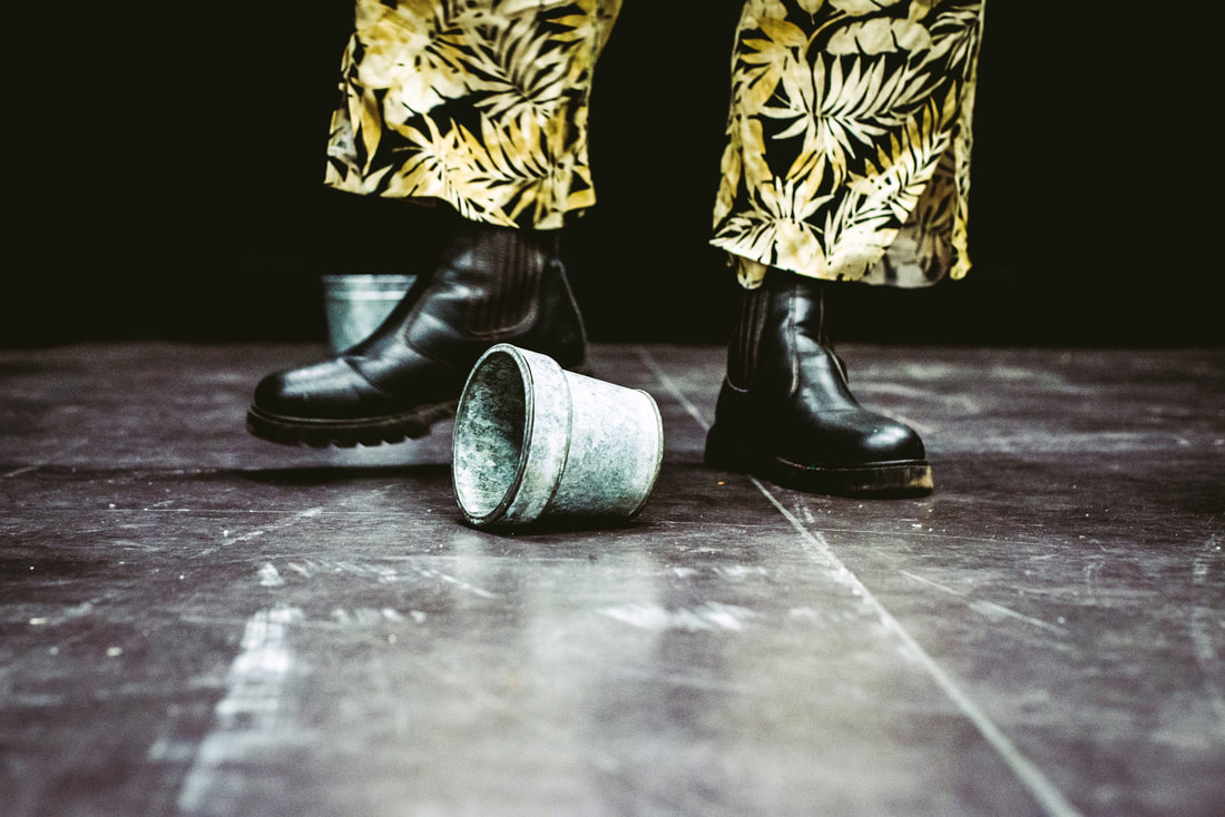Close up of gobscure’s feet wearing shiny black chunky boots, kicking a small silver bucket. He is wearing leaf print trousers which we can only see the bottom of.