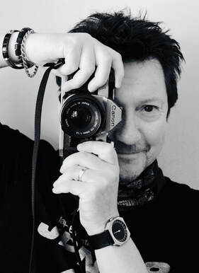 Black Robin, a white man in his fifties, in a black and white photograph. He has black spiky hair, bracelts on his right arm and is holding a vintage canon camera up to his eye. He is smiling.. 
