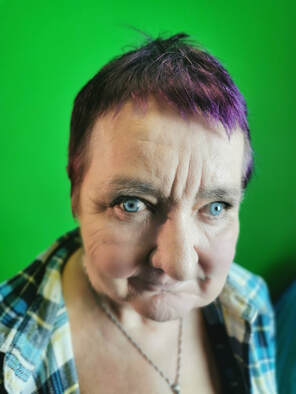 P)hoto of Karen Sheader, a woman in her late fifties with short plum dyed hair. She is wearing a blue and cream tartan shirt and is wearing a silver pendant. Karen is looking directly into the camera with a thoughtful smile. 