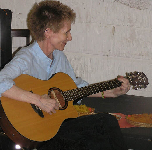 Image description - musician and songwriter Lindsay Carter pictured with guitar. Lindsay has short spiky hair, has a couple of earrings in the top of her right ear, and is wearing a light blue shirt and dark trousers. She is smiling whilst tuning her guitar. Pictured at Waygood Gallery for the launch of 21: The Last Avant Garde exhibition.