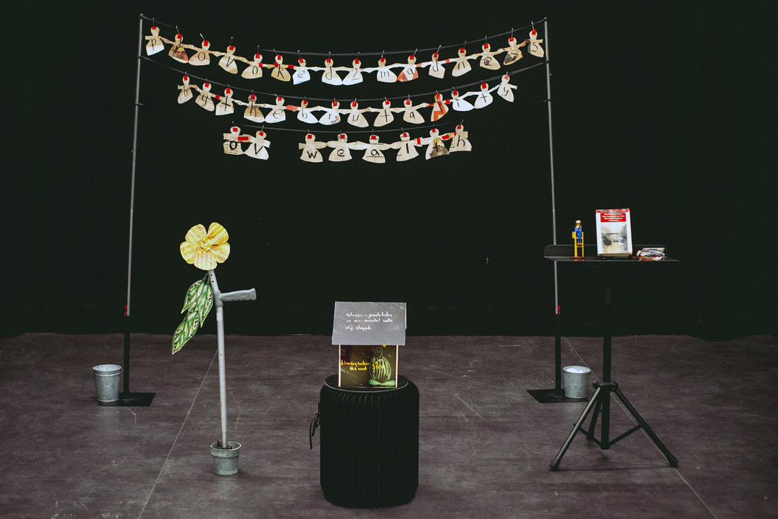 Photo of the set for 'Provoked to madness by the brutality of wealth', the words of which are written on several paper doll chains held on two poles at the back. 2 small silver buckets are at the base of each pole. A silver crutch in a plant pot with a large yellow flower and green leaves made from paper is stage left. Stage right is a small table with three small items on it. Centre stage is a small house with no walls and only a roof with a drawn figure seated underneath it. 
